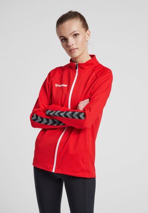 Jonge vrouw met haar naar achter gebonden, draagt een rode sportjas met ritssluiting en zwarte chevronstrepen op de mouwen, staat met gekruiste armen.