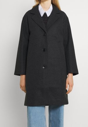 Lang mørkegrå knapjakke båret over en sort sweater og hvid skjorte med krave, kombineret med lyseblå jeans.