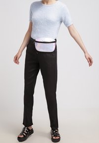 Marsupio realizzato in materiale iridescente con una zipper nera, indossato sopra un maglione blu di peluche a maniche corte e pantaloni neri, abbinato a sandali neri.