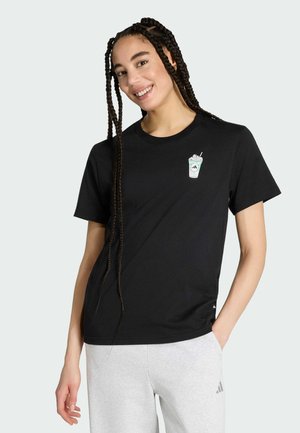 Junge Frau mit langen Zöpfen lächelt, trägt ein schwarzes T-Shirt mit einem kleinen Grafikdruck eines eisgekühlten Getränks und hellgraue Adidas-Sweatpants.