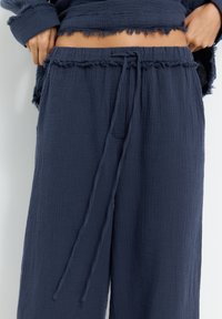 Navyblaue, strukturierte weit geschnittene Hosen mit einem ausfransenden Bund und einem Kordelzug. Der Stoff wirkt weich und leicht.