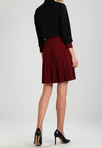 Chaqueta negra corta sobre un vestido burdeos con pliegues. Zapatos de tacón negro. Tejido suave y diseño estructurado con mangas hasta los codos.
