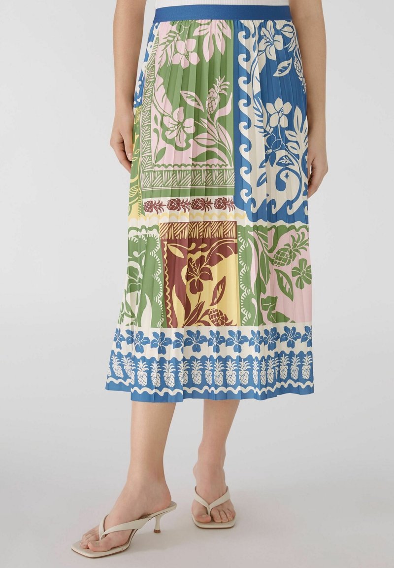 Jupe midi plissée avec imprimé patchwork floral multicolore et ananas, portée avec des sandales beiges à talons ouverts sur un fond clair.