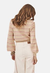 Suéter corto de estilo crochet en beige con rayas rosas y verdes, con mangas acampanadas. Combinado con jeans blancos de cintura alta.