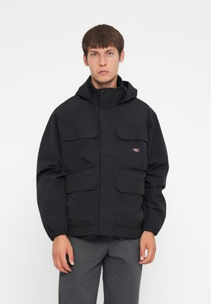 Black nylon jacket with a hood, featuring four front pockets, elastic cuffs, and a small logo on the left chest. The design is minimalistic.