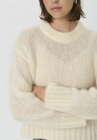Pulover kremne barve iz mehkega, prosojnega blaga z okroglim rebrastim izrezom in rebrastimi manšetami, s sproščenim krojem in vidnimi šivi.