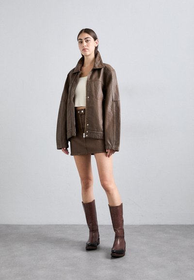 Brun læderjakke med en afslappet pasform, kombineret med en brun denimnederdel og knæhøje brune støvler, mod en neutral grå baggrund.