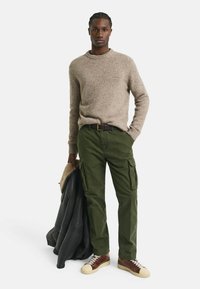 Beiger Strickpullover, olivgrüne Cargo-Hosen mit Seitentaschen, braune Leder-Sneaker mit cremefarbenen Sohlen, hält eine schwarze Jacke mit Pelzkragen.