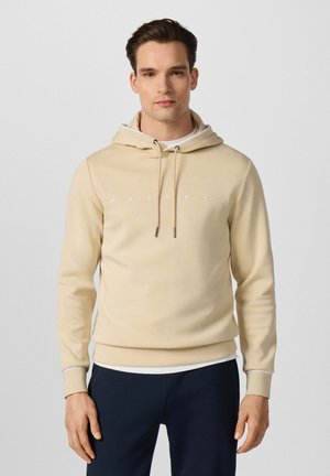 Man draagt een beige hoodie met trekkoorden en zwarte broek, staande tegen een effen achtergrond.