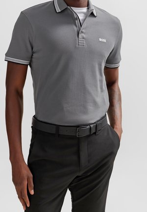 Homme portant un polo BOSS gris avec des bordures blanches, une ceinture noire et un pantalon noir, debout avec une main dans la poche devant un fond uni.