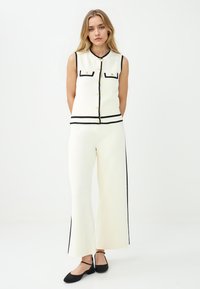 Creme-kleurige mouwloze top met zwarte biezen en twee voorzakken, gecombineerd met creme wijd uitlopende broek met zijdelingse zwarte strepen.