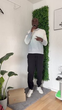 Person wearing a light gray-to-white gradient sweater and black pants, standing on a beige mat near a green plant and a cardboard box.