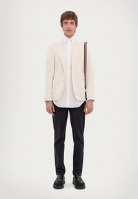 Cremefarbener Blazer, weißes Hemd, dunkle Hose und schwarze Schuhe. Der Blazer zeichnet sich durch ein schlankes Design mit subtiler Textur und zwei Fronttaschen aus.