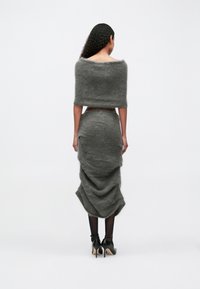 Ženska z dolgimi kodrastimi lasmi, oblečena v puhast sivi top brez naramnic in usklajen, naguban midi krilo z črnimi nogavicami in petkami, stoji obrnjena stran od kamere.