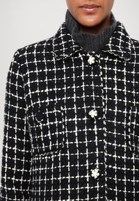 Chaqueta de tweed en blanco y negro con un diseño a cuadros, cuello, cierre de botones con acentos de flores y bolsillos delanteros. Tejido texturizado.