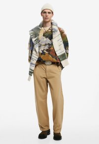 Maglione in maglia grossa con un motivo paesaggistico multicolore, pantaloni beige e calzature scure. Gli accessori includono un berretto in maglia e una cintura.