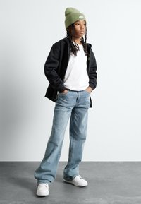Črna zip-jacket, bela majica, svetlo modri jeans, zeleni zimski klobuk in beli superge. Model stoji z rokami v žepih na sivi tla.