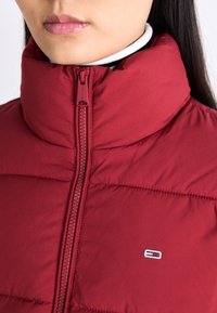 Zbliżenie na osobę w czerwonej kurtce puchowej z wysokim kołnierzem i małym prostokątnym logo na klatce piersiowej.