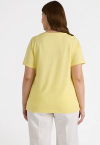 Camiseta amarilla de manga corta vista desde la espalda, con un cuello redondo y una textura suave y lisa. Combinada con pantalones de color claro.