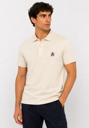 Homme portant un polo beige à manches courtes avec un petit logo sur la poitrine et un pantalon bleu foncé, debout avec une main dans la poche.