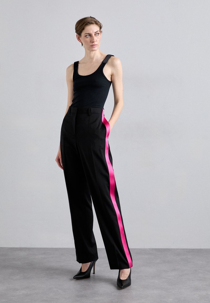 Pantaloni neri su misura con strisce laterali in satin rosa brillante, abbinati a una canotta nera aderente. Tacchi neri con fasce completano il look.