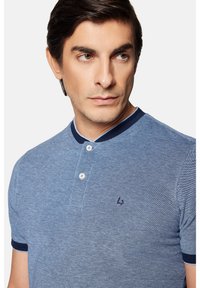 Niebieska koszulka polo z teksturowanym wzorem, dwa guziki przy kołnierzyku oraz granatowe wykończenia na kołnierzu i rękawach. Zawiera małe logo na piersi.