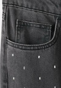Tejido de mezclilla negra que presenta un bolsillo trasero adornado con pequeños remaches rectangulares plateados dispuestos en un patrón disperso.