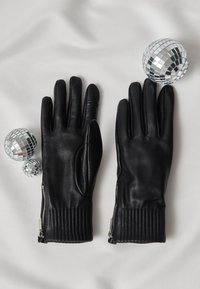 Schwarze Lederhandschuhe mit gerippten Strickbündchen, versehen mit Reißverschlüssen am Handgelenk. Begleitet von zwei spiegelverkehrten Diskokugeln vor hellem Hintergrund.