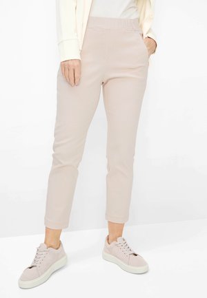 Kvinde iført lyse beige, strukturerede, figursyede bukser, cremefarvede sneakers og en cremefarvet langærmet top, stående med den ene hånd i lommen.