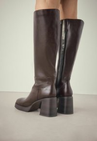 Brązowe skórzane kozaki do kolan z zaokrąglonym noskiem, masywnym obcasem i bocznym zamkiem błyskawicznym. Gładka faktura z eleganckim designem.