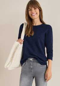 Marineblaues Strickoberteil mit Dreiviertelärmeln, kombiniert mit hellgrauen Jeans und einer strukturierten cremefarbenen Tragetasche.