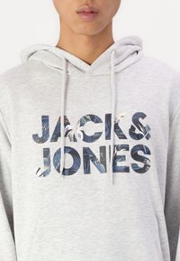 Grauer Kapuzenpullover mit einer Vorderfronttasche, auf der der Markenname "JACK & JONES" in fetten Buchstaben mit einem Blumenmuster in Blau und Grün gedruckt ist.