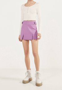 Personne portant un crop top blanc à manches longues, une mini-jupe violette avec un détail à double boucle, et des bottines blanches à lacets avec des semelles épaisses.