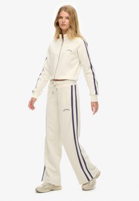 Beige zip-up jacket en bijpassende broek met blauwe strepen. Katoenmix stof, relaxte pasvorm en logo detail op de borst en dij.