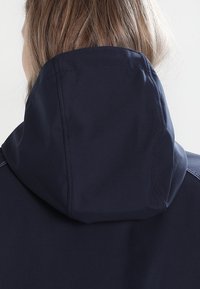 Blond person sedd bakifrån iförd en marinblå huvtröja med synliga sömmar mot en ljus bakgrund.