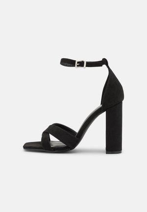 Sandale noire en daim à talon haut avec bride à la cheville, boucle argentée, talon bloc et brides croisées à l’avant sur un modèle à bout ouvert.