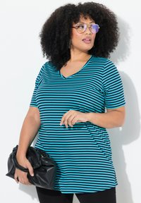 Camiseta de rayas en verde azulado y negro, con escote en V, mangas cortas y un ajuste holgado. El modelo sostiene un bolso de mano negro.