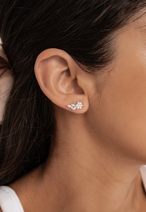 Nahaufnahme eines Frauenohres, das einen silbernen Blütenohrring mit mehreren kleinen, funkelnden Steinen trägt.
