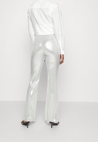 Person wearing shiny silver flared pants, white long-sleeve shirt, and black high-heeled sandals, standing against a plain background.