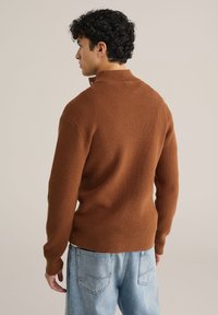 Rudas megztas svarkas su aukšta apykakle, raukšlėtomis rankogaliais ir apačia. dėvimas su šviesiai mėlynomis džinsinėmis kelnėmis, matomas iš nugaros.