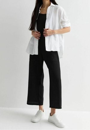Femme portant une chemise blanche boutonnée par-dessus une combi-pantalon noire sans manches et des baskets blanches, debout devant un fond clair uni.
