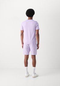 Lilla kortermet t-skjorte og matchende shorts. Slitesterk tekstur, enkelt design. Stylet med hvite sokker og joggesko, sett fra bak.
