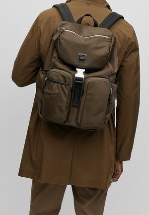 Osoba v hnědém saku a kalhotách s velkým hnědým designovým batůžkem, který má několik zipových kapes a přední sponu.