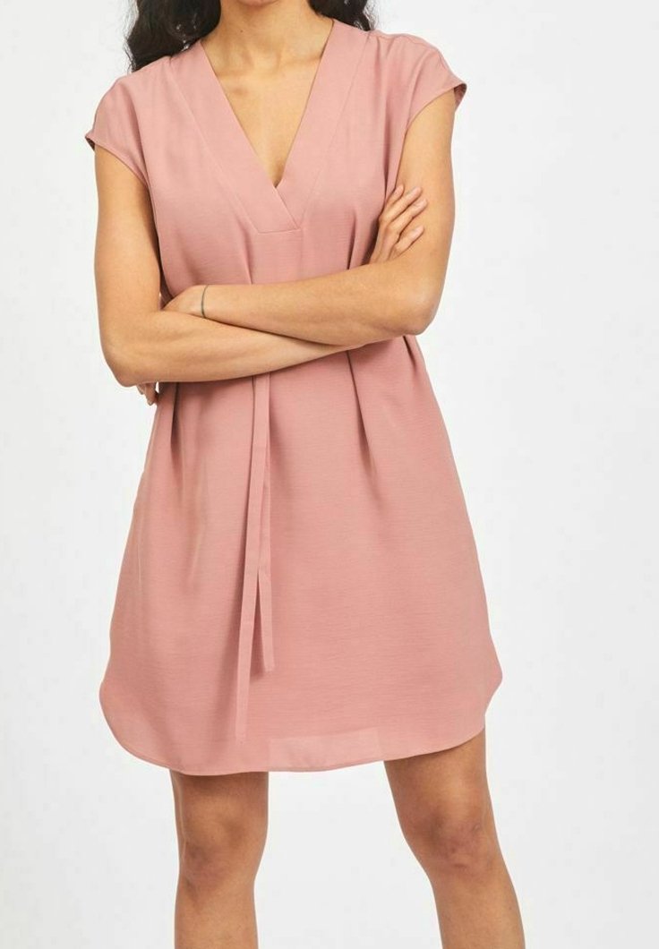 Femme portant une robe courte à manches courtes, rose poudré, longueur genou, avec un décolleté en V et les bras croisés sur la poitrine sur un fond uni.