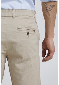 Pantalones beige con una textura suave, que presentan un bolsillo trasero con un botón marrón y un acento a rayas. Primer plano de la cintura y la mano.