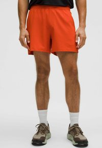 Mann in leuchtend orangen Sport Shorts, weißen Socken und braunen sowie beigen Laufschuhen, der mit leicht gespreizten Beinen steht.