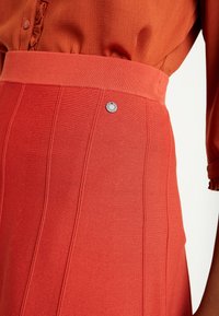 Jupe en maille rouge à texture côtelée, avec une large ceinture. Détail argenté attaché à la ceinture. Coordonnée avec un haut boutonné.