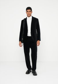 Completo in velvet nero con giacca slim-fit e pantaloni. Camicia bianca sotto. Scarpe nere e borsa a tracolla completano il look.