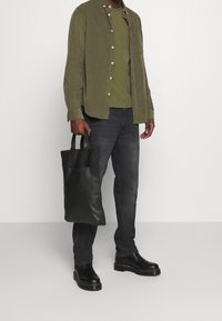 Černá kožená taška přes rameno s krátkými uchy, olivově zelená manšestrová košile, zelené tričko pod ní, tmavé džíny a černé boty.