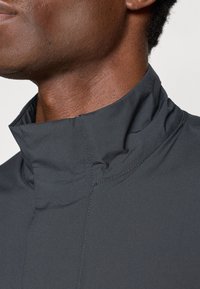 Veste noire à col haut avec une texture lisse et des détails cousus. Présente un décolleté net et un design moderne sans fermetures.